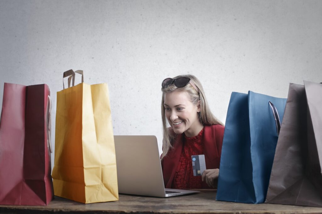 Mulher olhando para a tela do computador e fazendo uma compra online rodeada de bolsas de compras coloridas